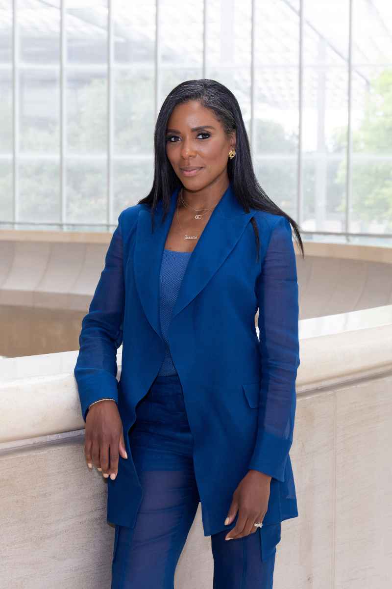A woman in a vibrant blue pantsuit smiles while leaning against a stone railing indoors.