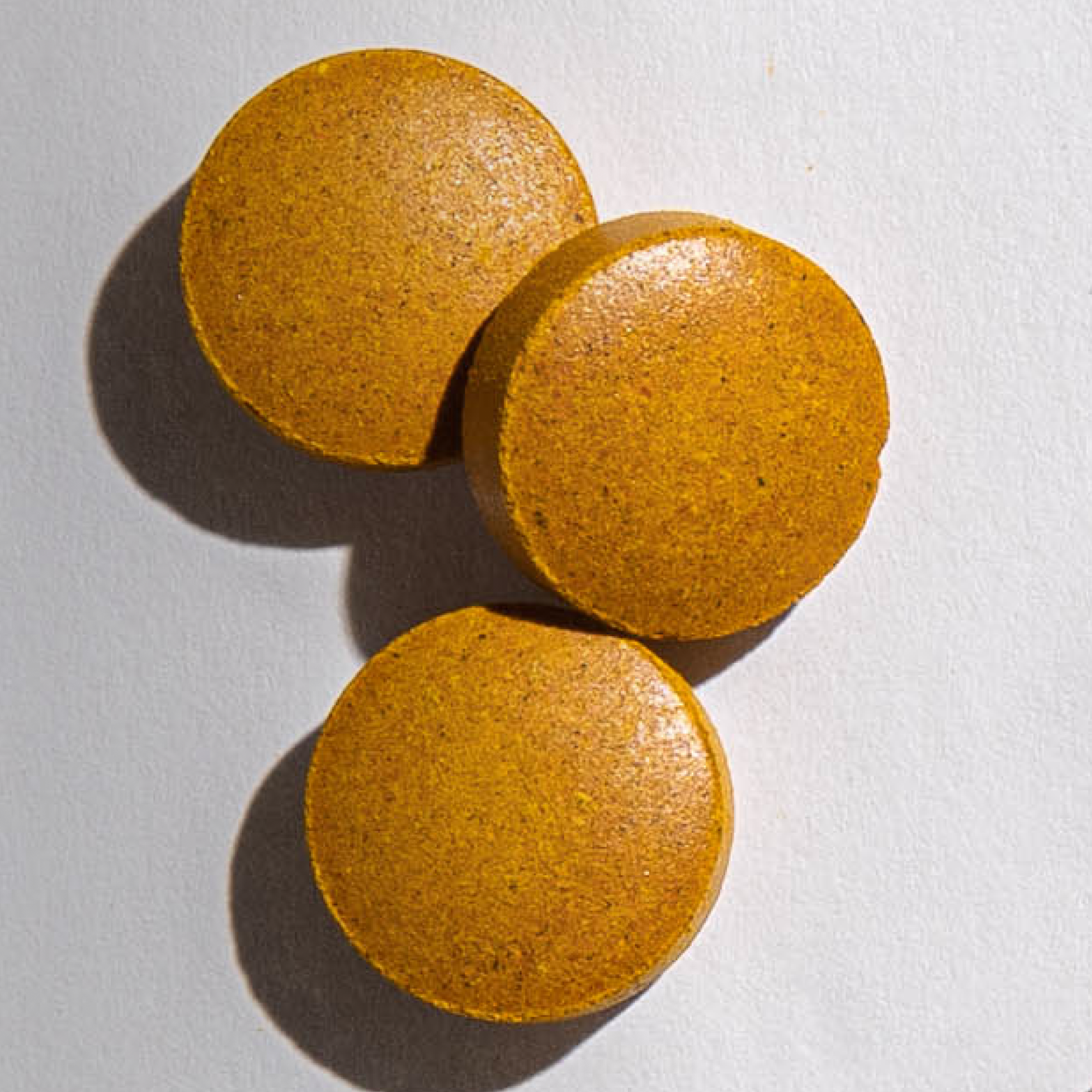 An overhead close-up of three round, speckled, golden-yellow tablets on a white surface.