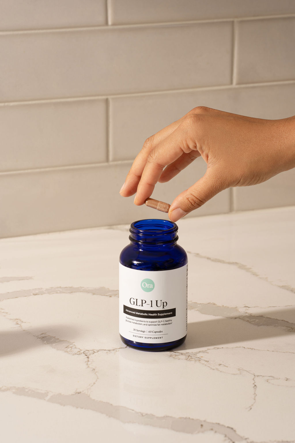 Hand holding a capsule above a blue supplement bottle on a marble surface.