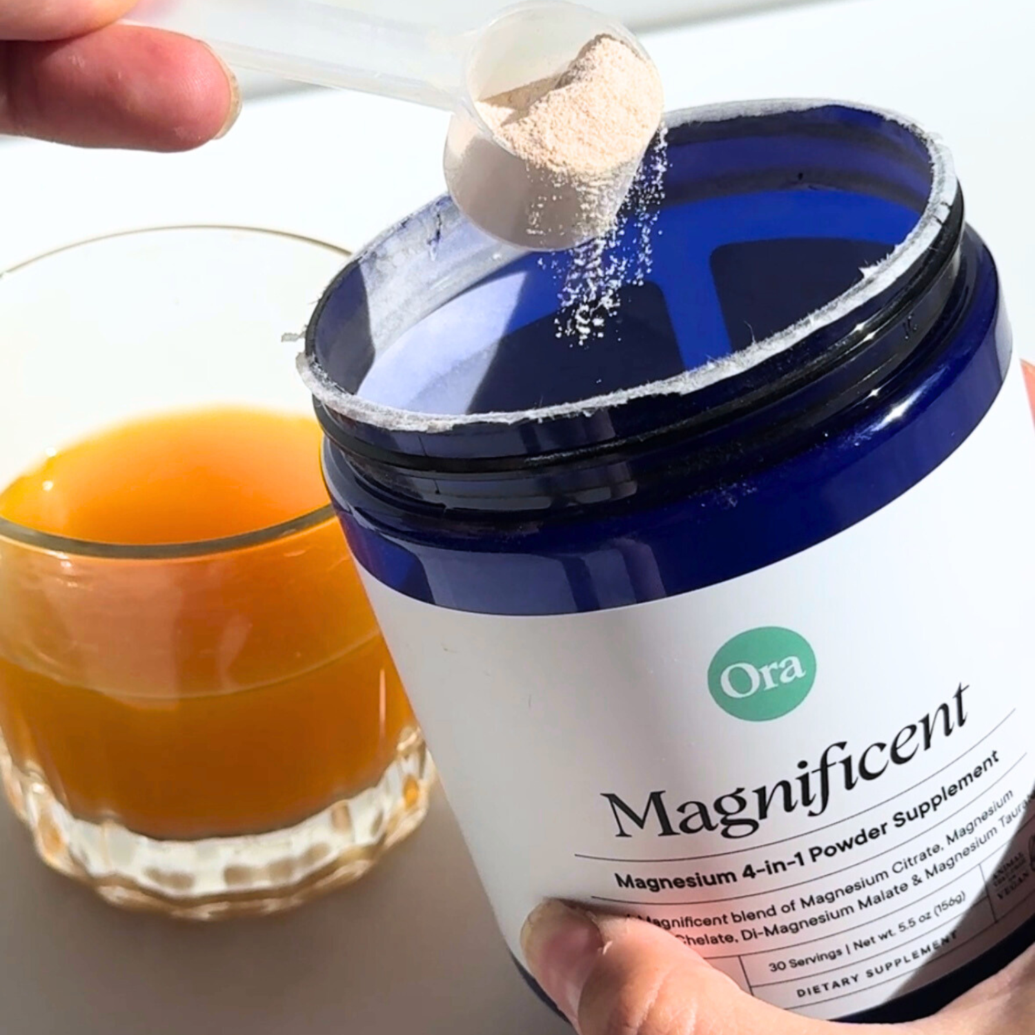 Scoop of powder being added to a jar labeled 'Magnificent' beside a glass of orange liquid.