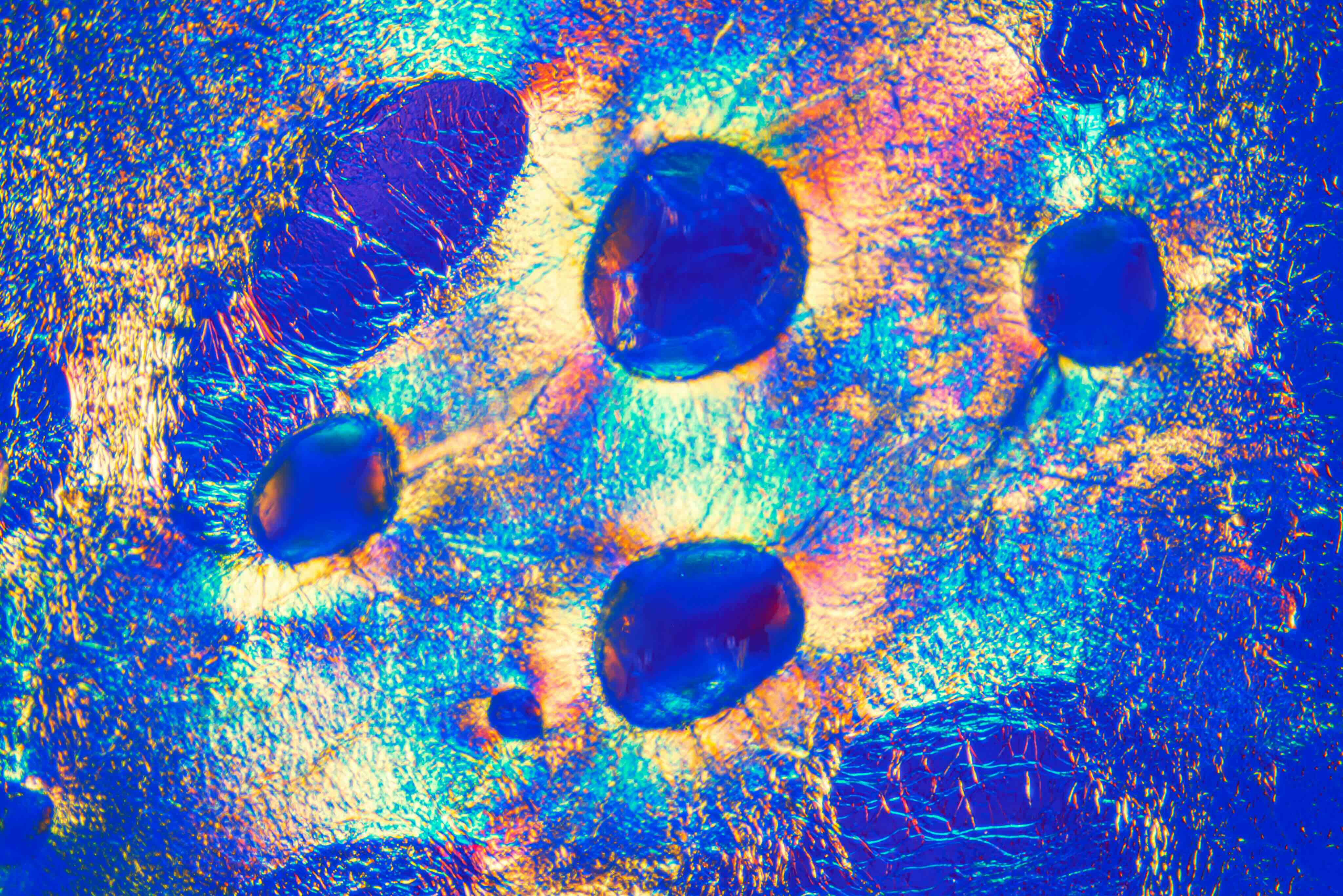 An abstract macro photo of a crinkled, iridescent surface in blue and gold with dark ovoid shapes.