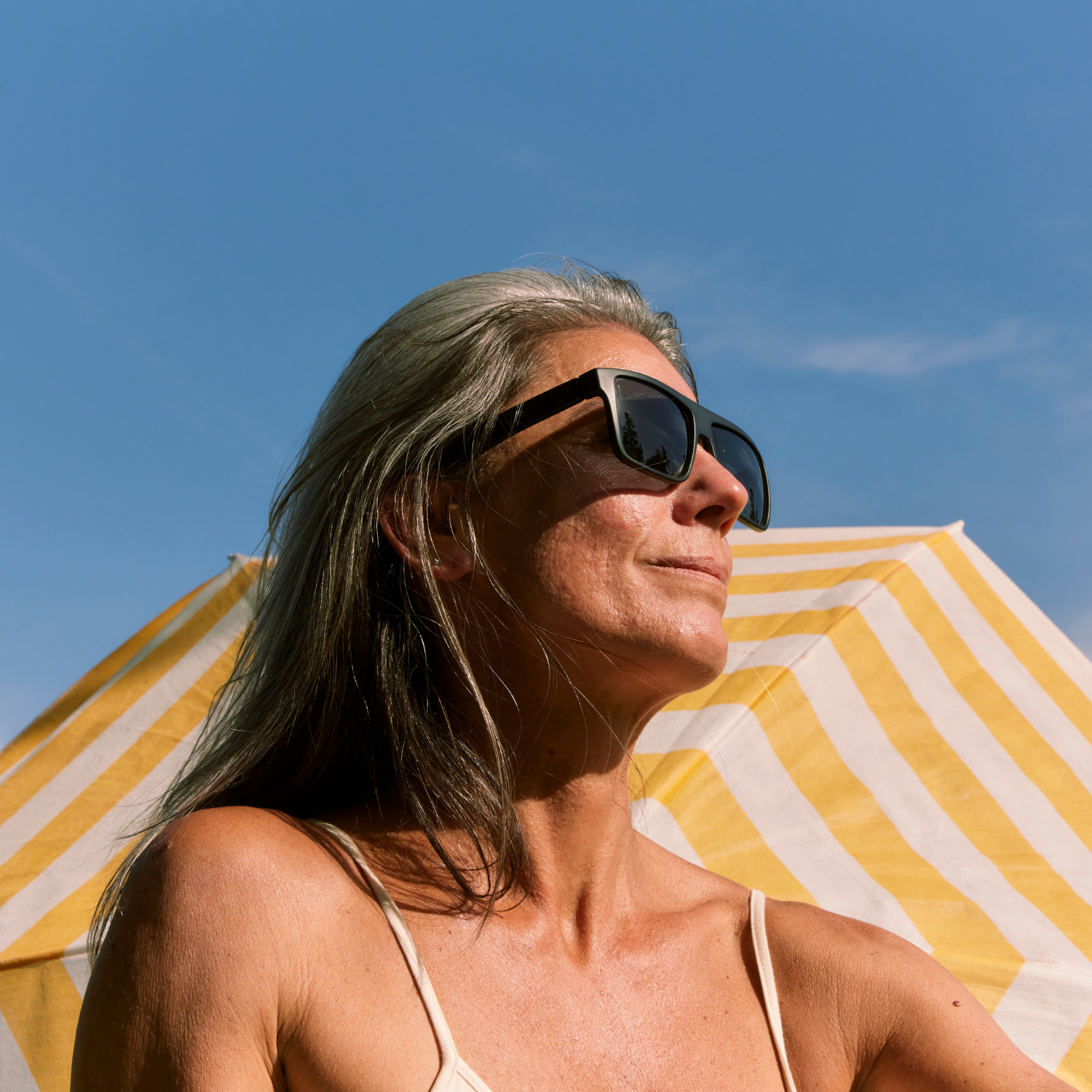 Person wearing sunglasses, sitting under a yellow striped umbrella, with a clear blue sky.