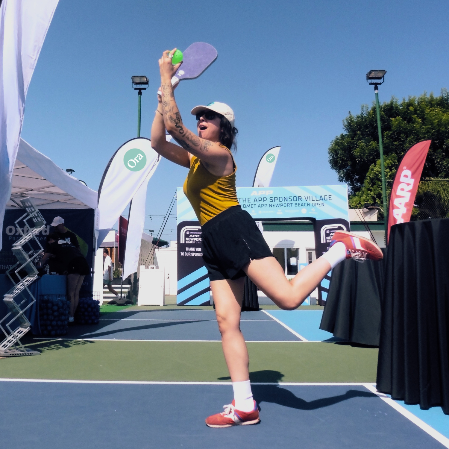 Person playing pickleball on a court, holding a paddle and ball, with sponsor banners in background.