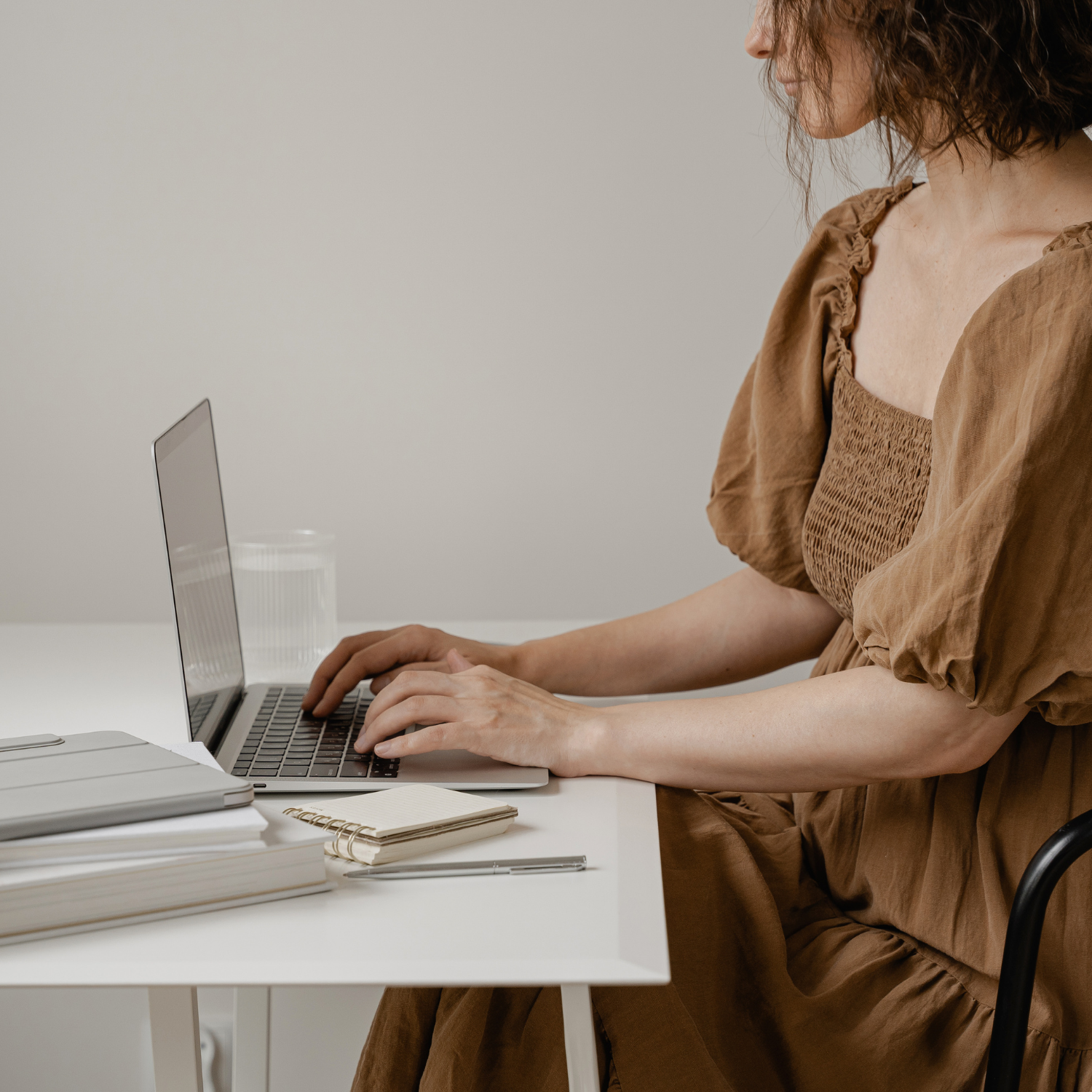 A woman in a brown dress sits at a white desk typing on a laptop.