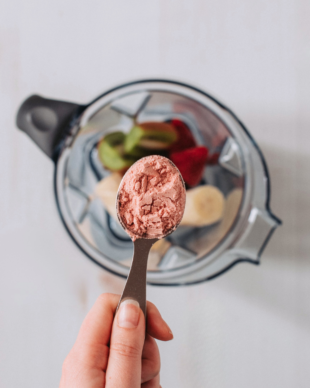 A hand holds a measuring spoon with pink powder over a blender containing fruit.