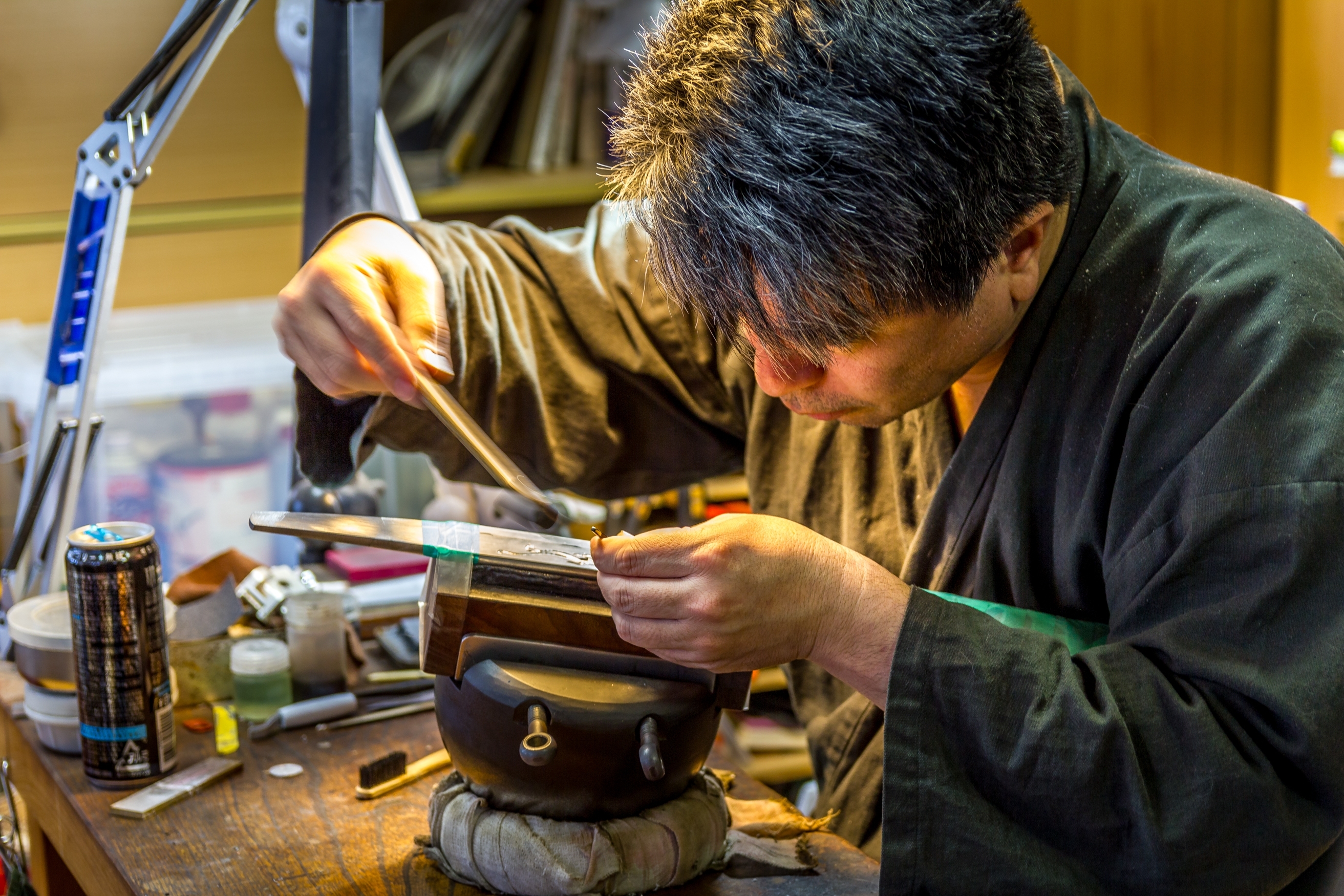 Person carefully working on a metal object at a cluttered workbench.