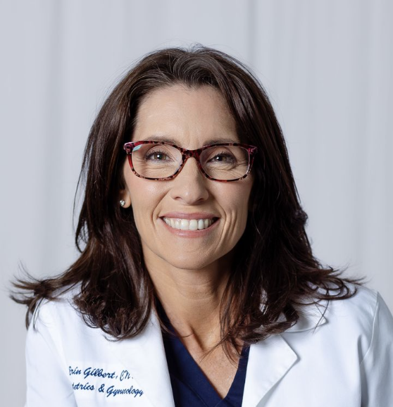 A headshot of a smiling woman with brown hair and glasses, wearing a white lab coat.