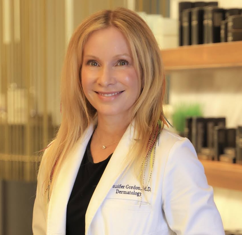 A smiling woman with blonde hair, wearing a white lab coat with 'Jennifer Gordon, M.D. Dermatology' embroidered on it.