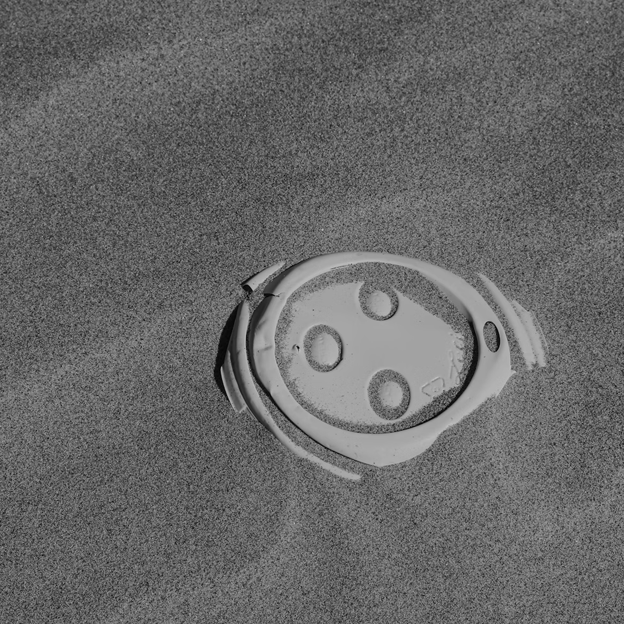 A black and white photo of a broken plastic lid partially buried in the sand.
