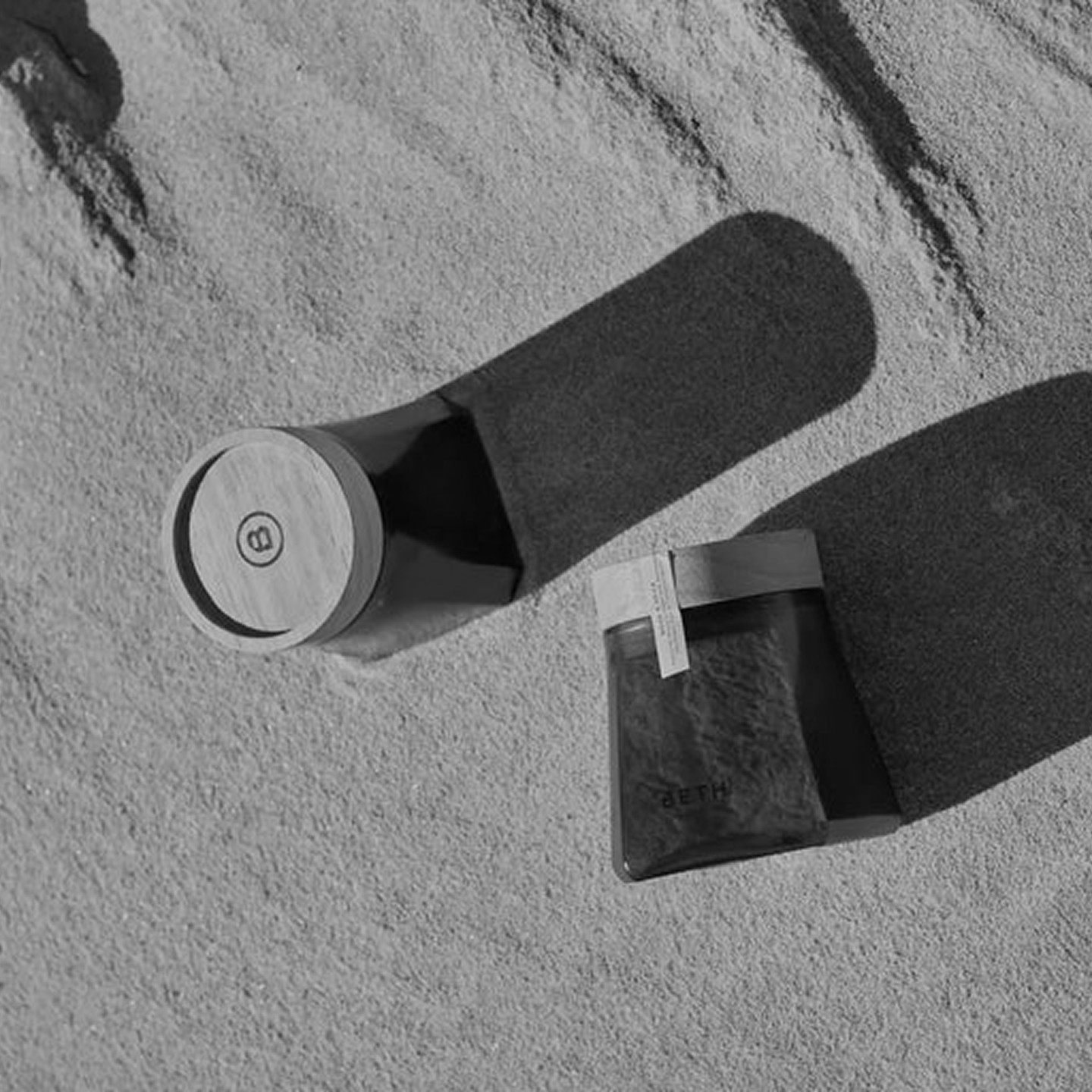 A black and white overhead photo of two jars with wooden lids on sand, casting long shadows.