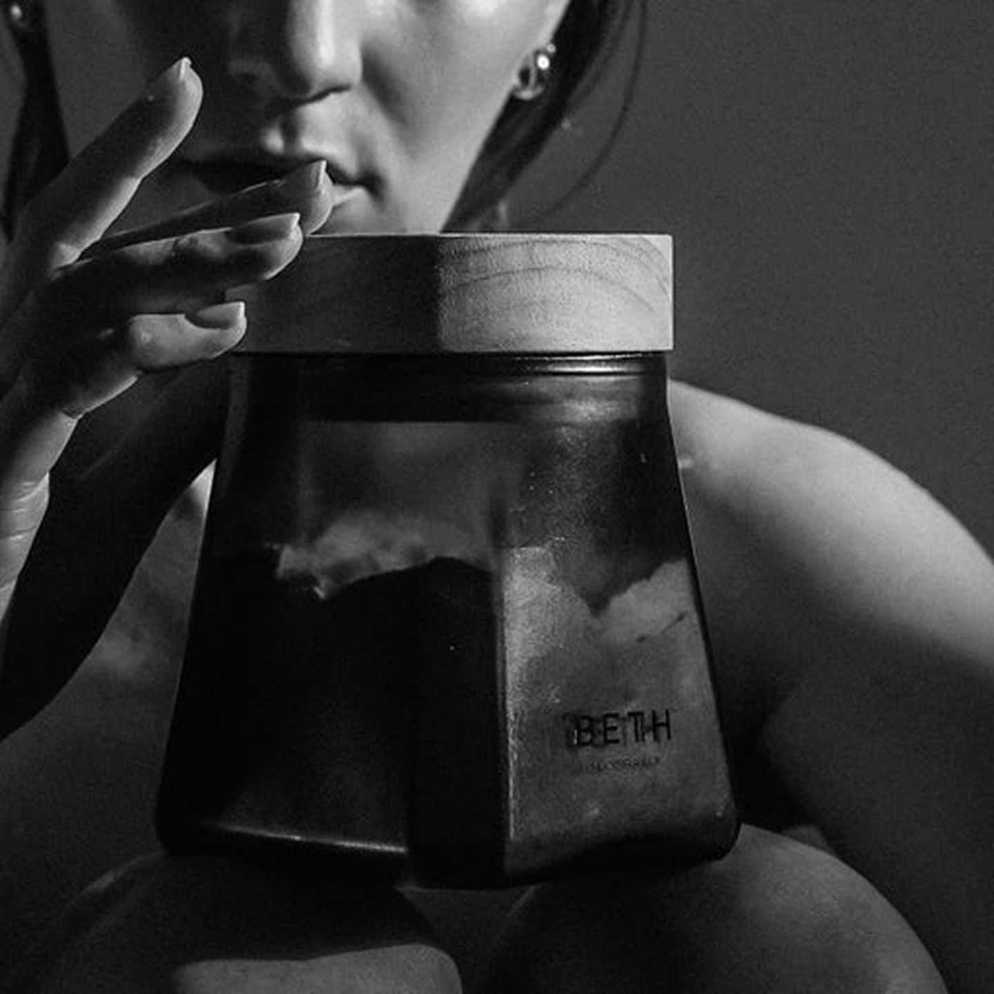 A close-up, black and white photo of a woman holding a dark jar with a wooden lid.