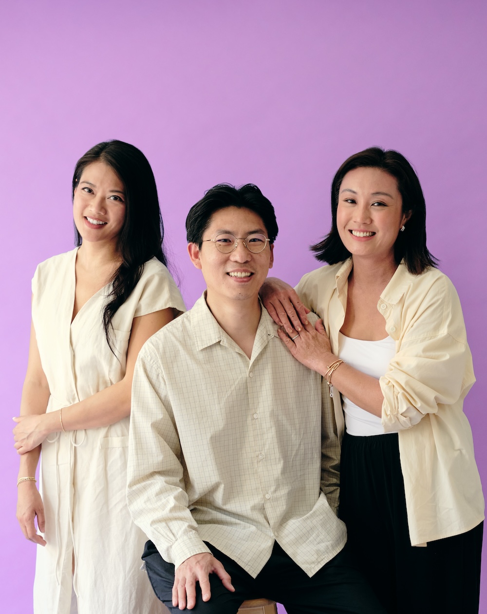 Two women and a man pose together smiling in front of a solid purple background.