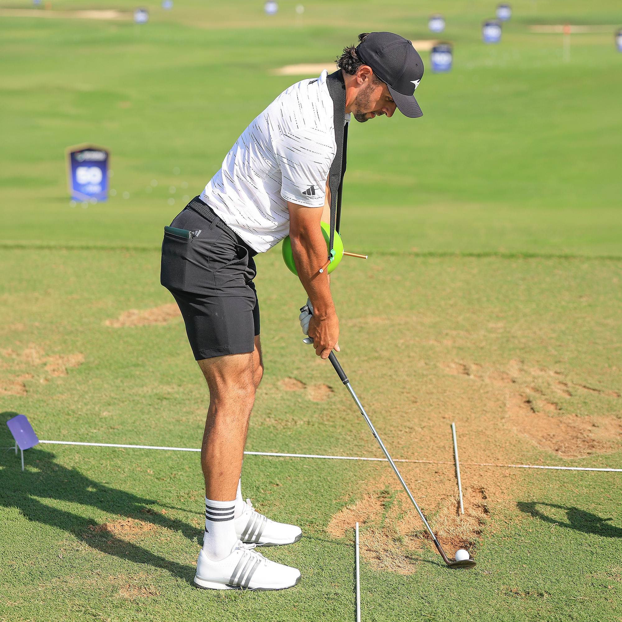 A male golfer on a driving range prepares to hit a golf ball while wearing a green training aid.