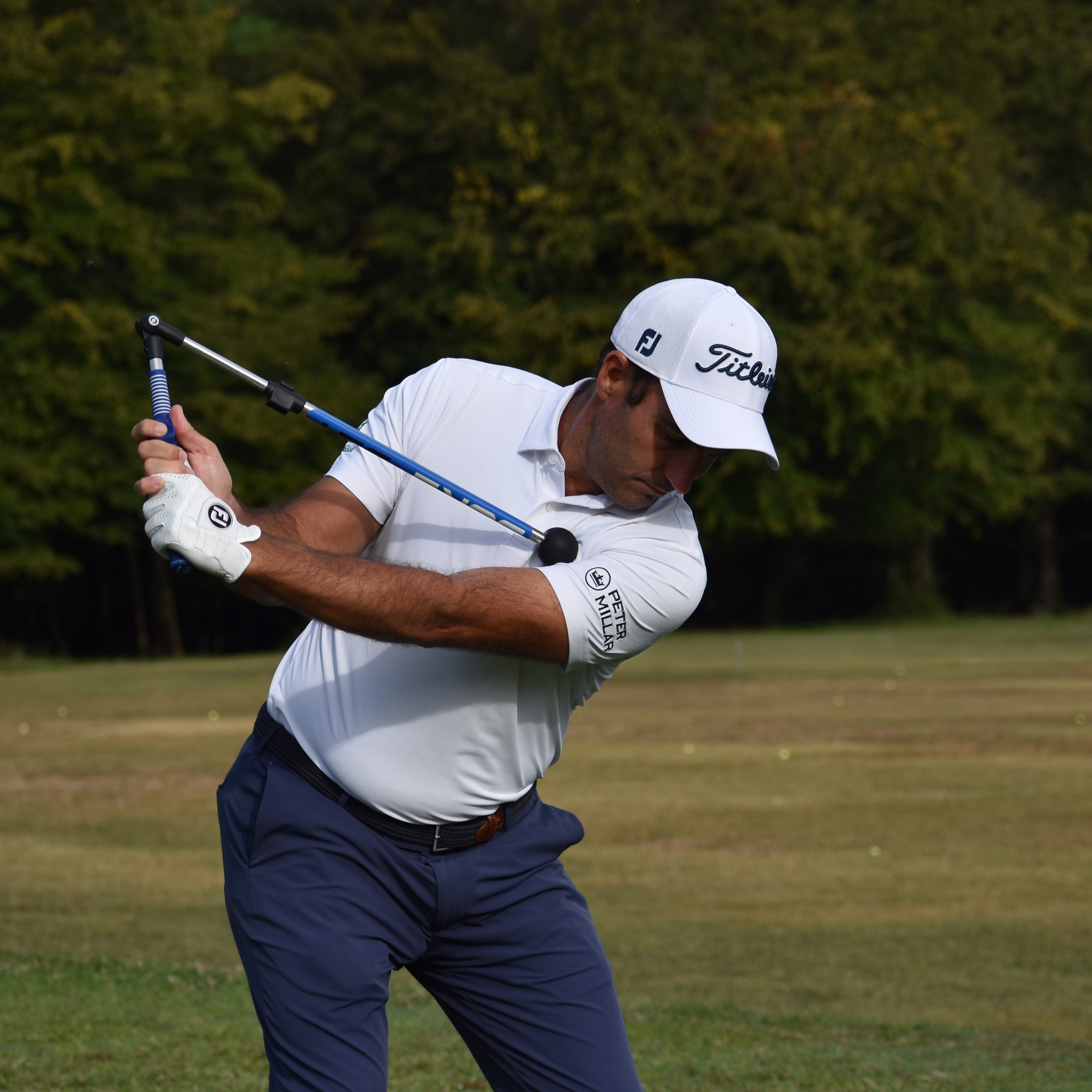 A male golfer in a white shirt and cap practices his swing with a blue training aid.