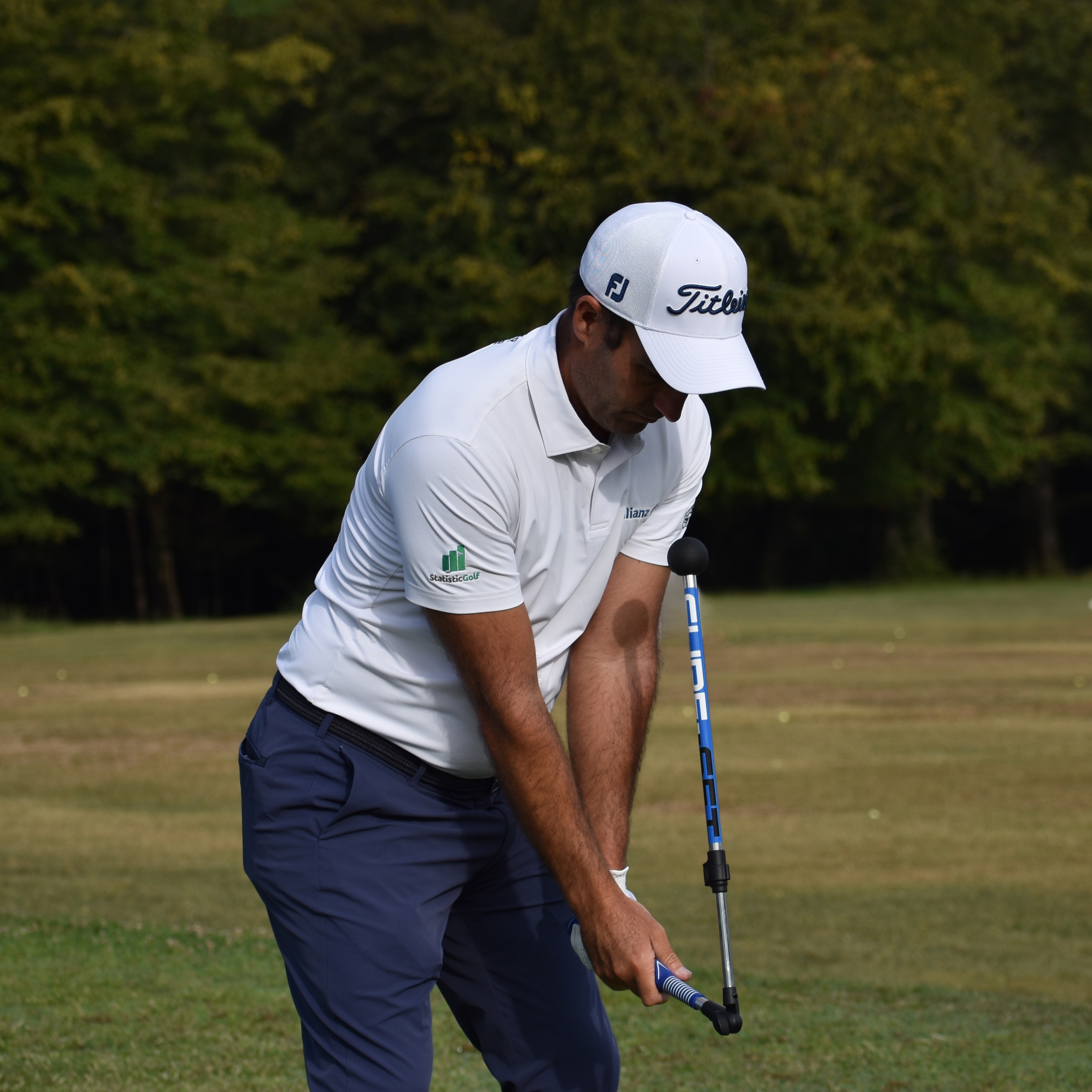 A male golfer in a white shirt and cap practices his swing with a blue training aid.