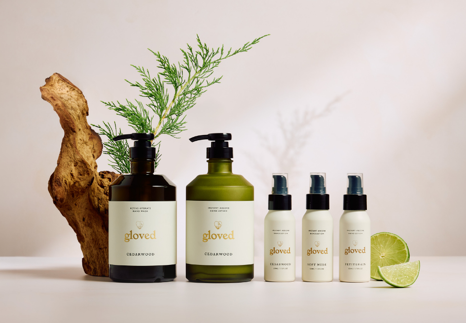 A collection of 'gloved' hand care products arranged with a cedar sprig, wood, and a sliced lime.