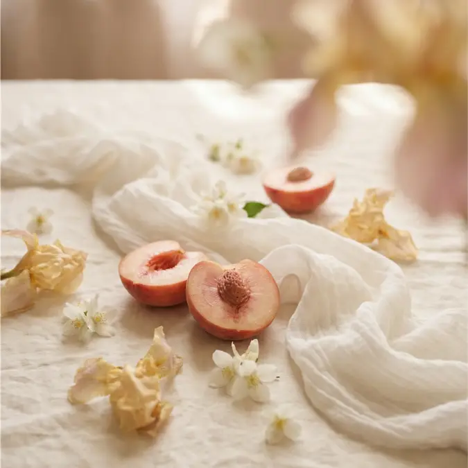 A still life of halved peaches and small white and yellow flowers on a rumpled white cloth.