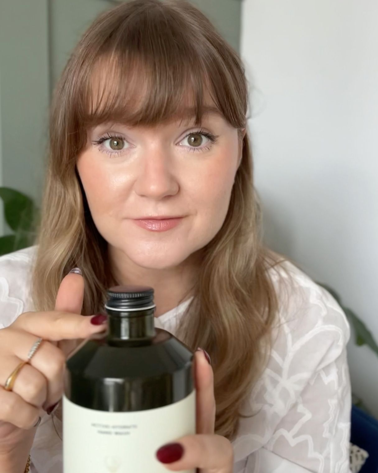 A woman with bangs and light brown hair holds a bottle, pointing to its cap while looking forward.