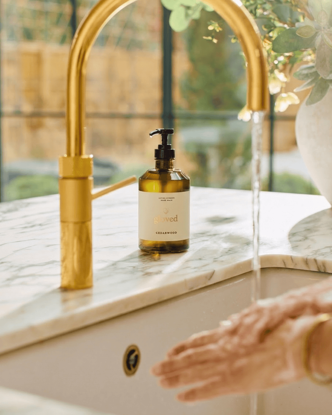 A person washes their hands in a sink with a gold faucet and a bottle of soap nearby.