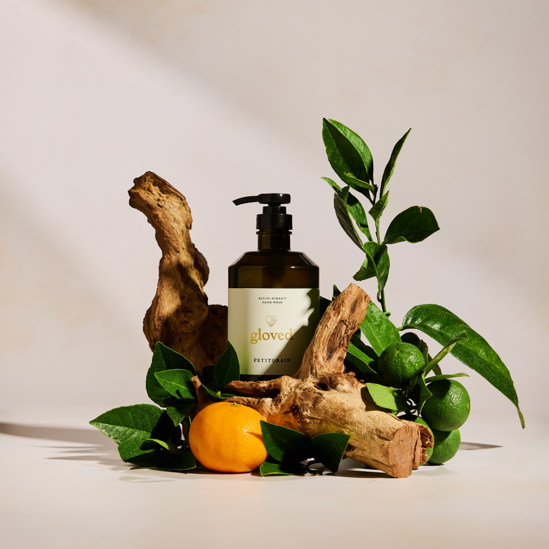 A bottle of Gloved hand wash arranged with driftwood, a tangerine, and a branch of limes.