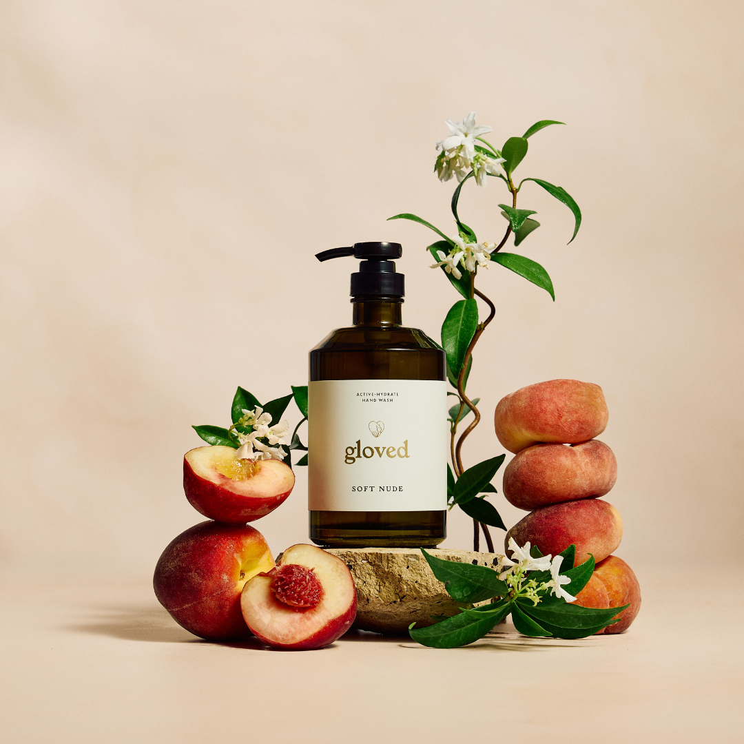 A bottle of Gloved hand wash sits on a stone, surrounded by fresh peaches and a jasmine vine.