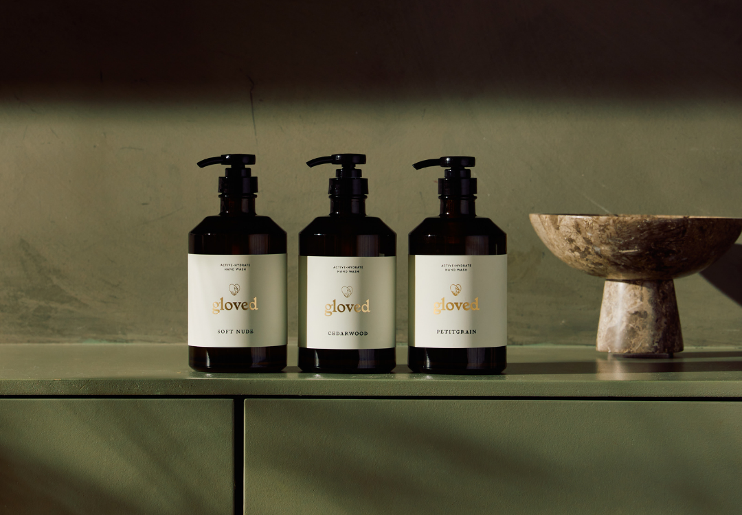 Three black pump bottles of 'gloved' hand wash sit on a green shelf next to a marble bowl.