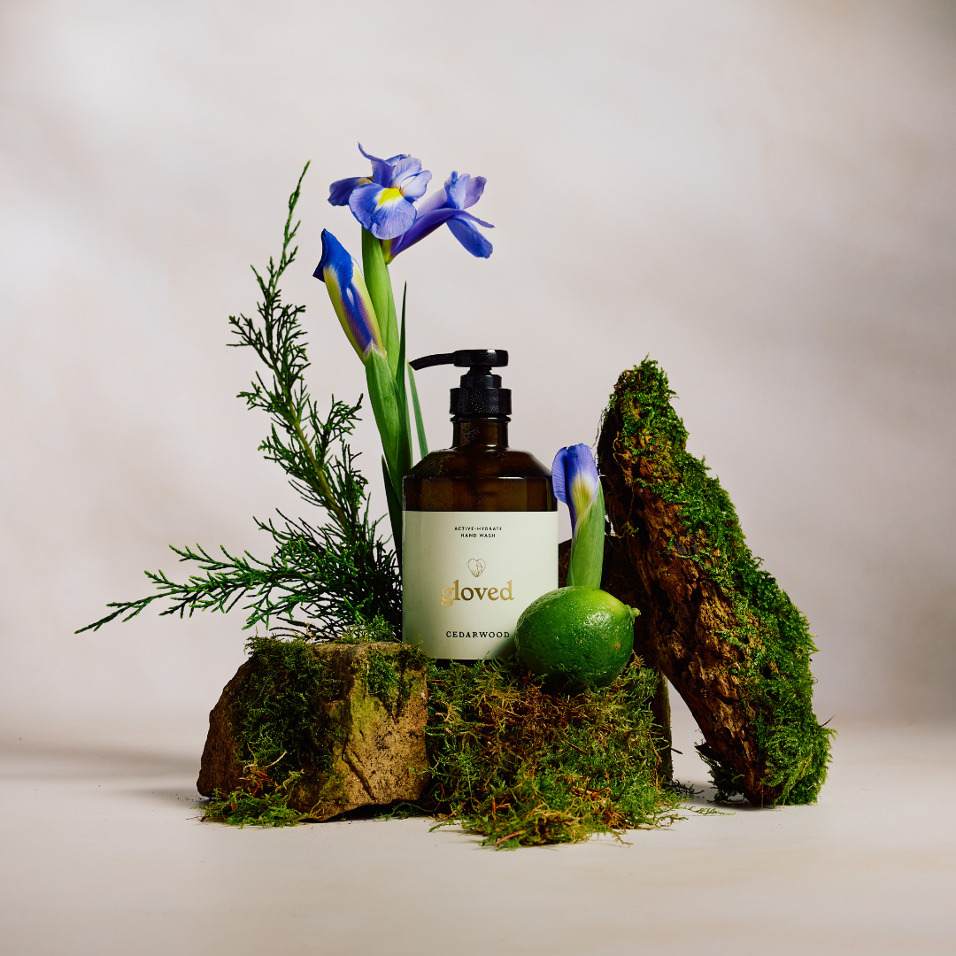 A bottle of Gloved Cedarwood hand wash sits among moss, purple irises, a lime, and an evergreen sprig.