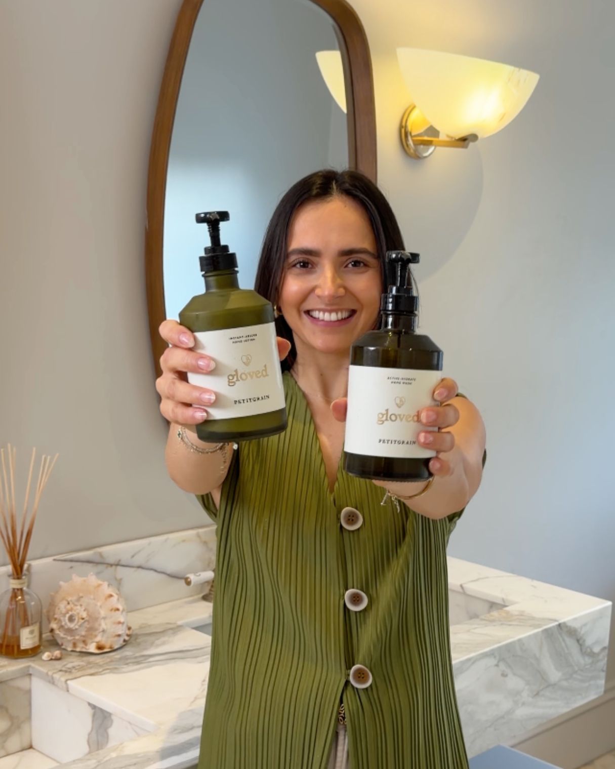 A smiling woman in a green top holds up two pump bottles in a bathroom.