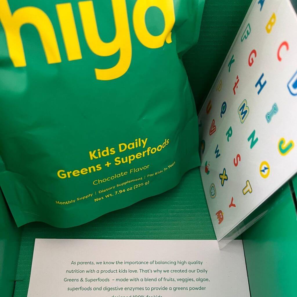 A green pouch of Hiya Kids Daily Greens + Superfoods next to a sheet of alphabet stickers.