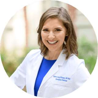A smiling woman with brown hair, wearing a white medical coat over a blue shirt.