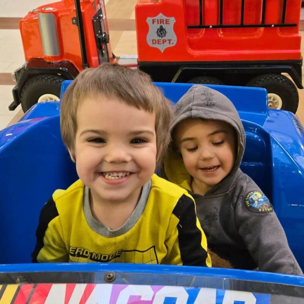 Two young children smile while sitting inside a blue toy car ride next to a toy fire truck.