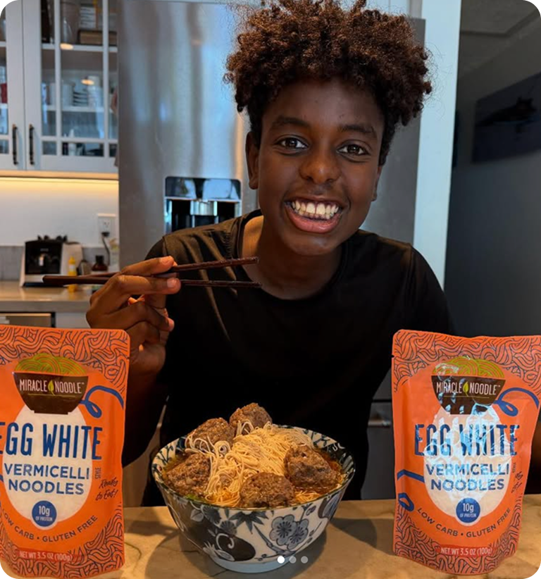 Person smiling, holding chopsticks with bowl of noodles and packages of vermicelli on the table.