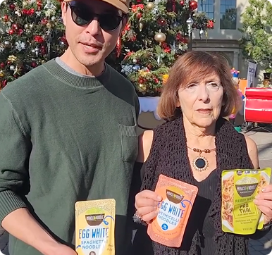 Two people holding packaged food products in front of a decorated Christmas tree.