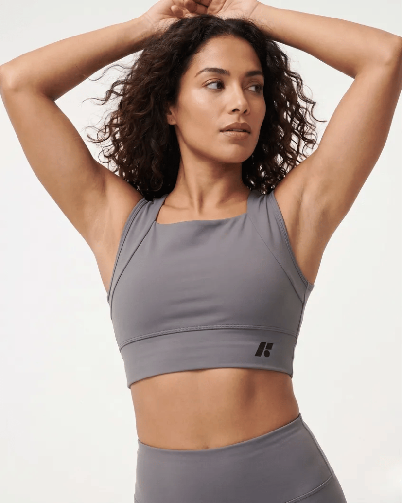 A woman with curly brown hair wearing a grey sports bra with her hands behind her head.