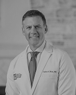 A black and white professional headshot of a smiling man wearing a white lab coat and tie.
