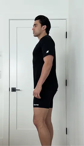 A man stands in profile wearing a black t-shirt and shorts, with his shoulders slumped forward.