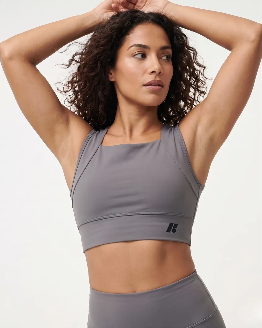 A woman in a grey sports bra and leggings poses with her hands behind her head.