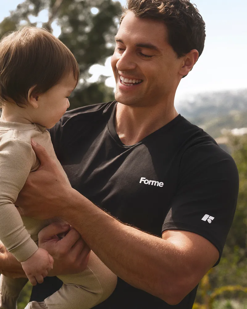 A smiling man wearing a black t-shirt with the word 'Forme' on it holds a small child.