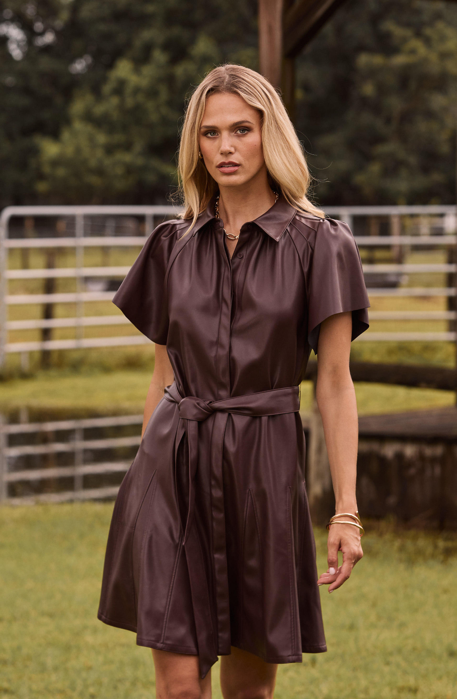 A person in a brown belted dress standing outside near a fence.