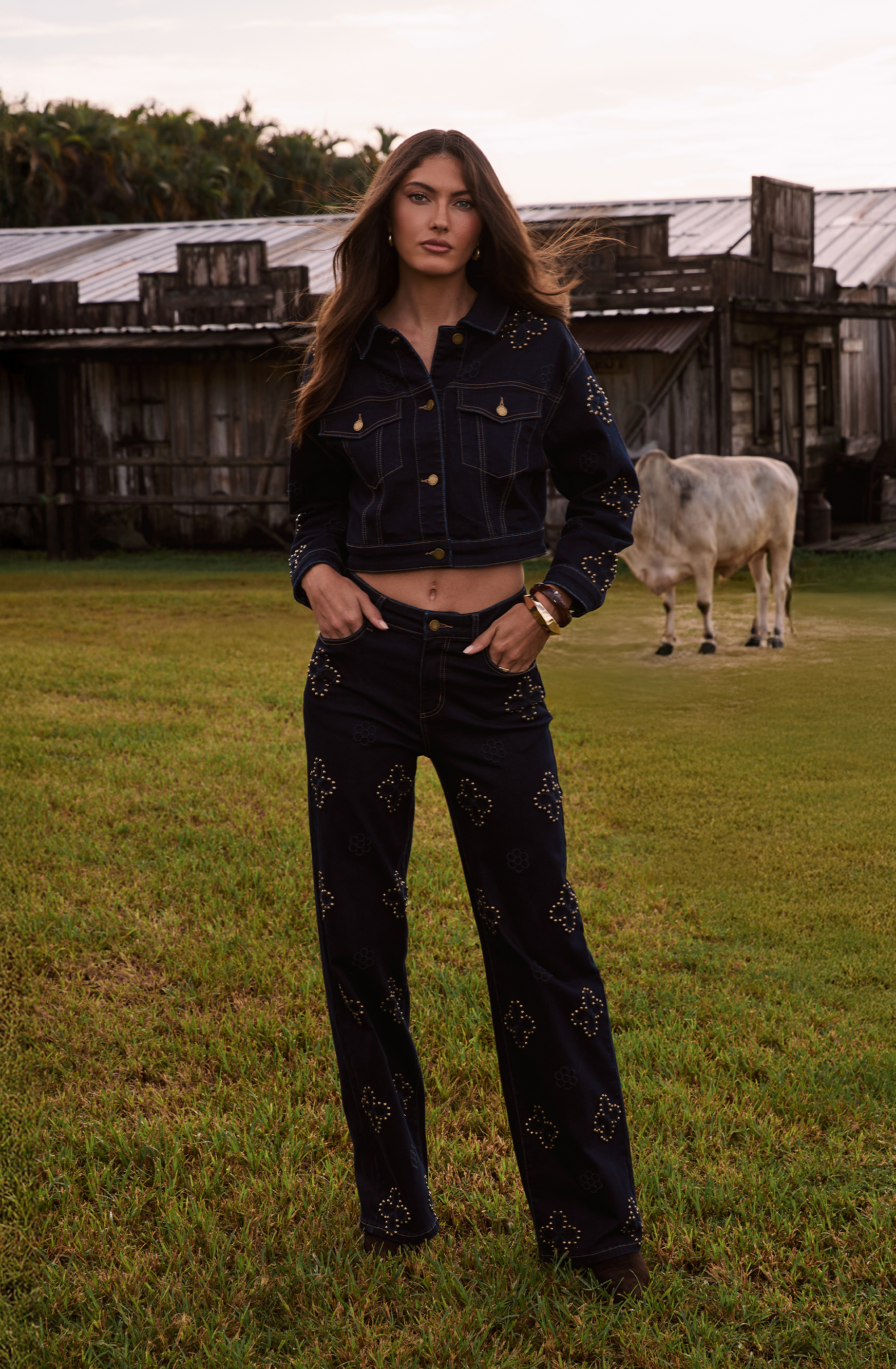 Person in denim outfit standing on grassy field with a cow in the background.