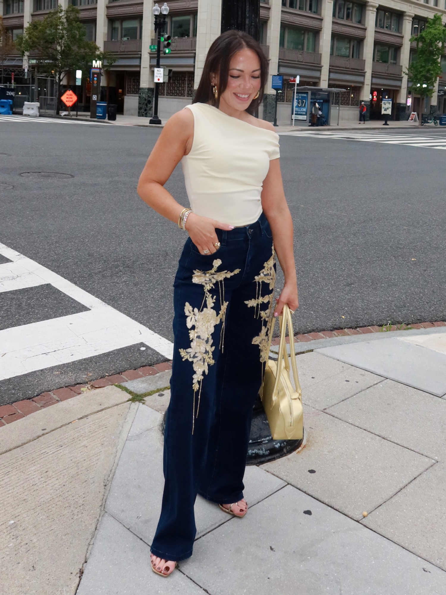 Person in white top and embroidered jeans standing on city sidewalk.