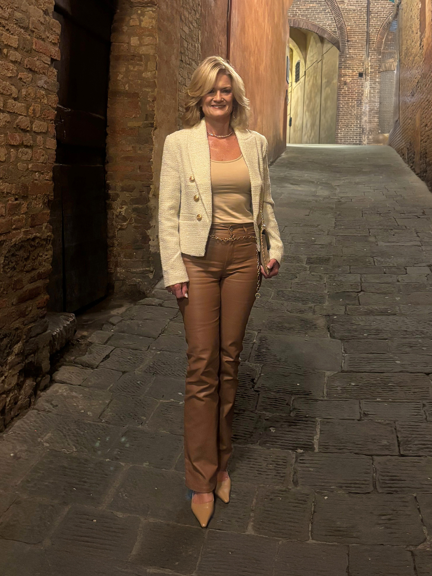 Woman in a beige outfit standing in a cobblestone alley with brick walls.