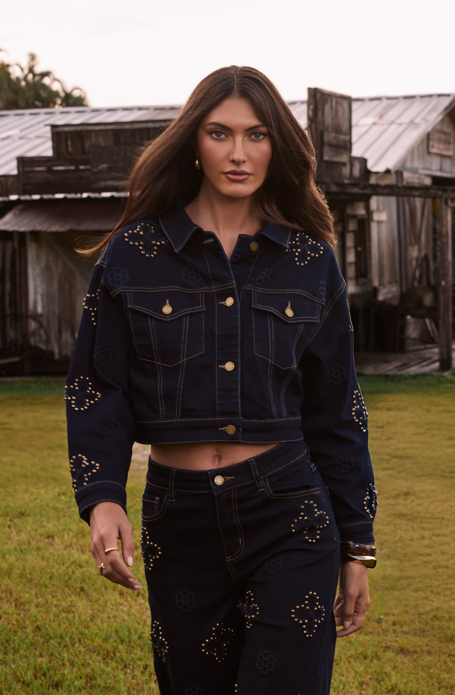 Person in a studded denim outfit standing outdoors near rustic buildings.