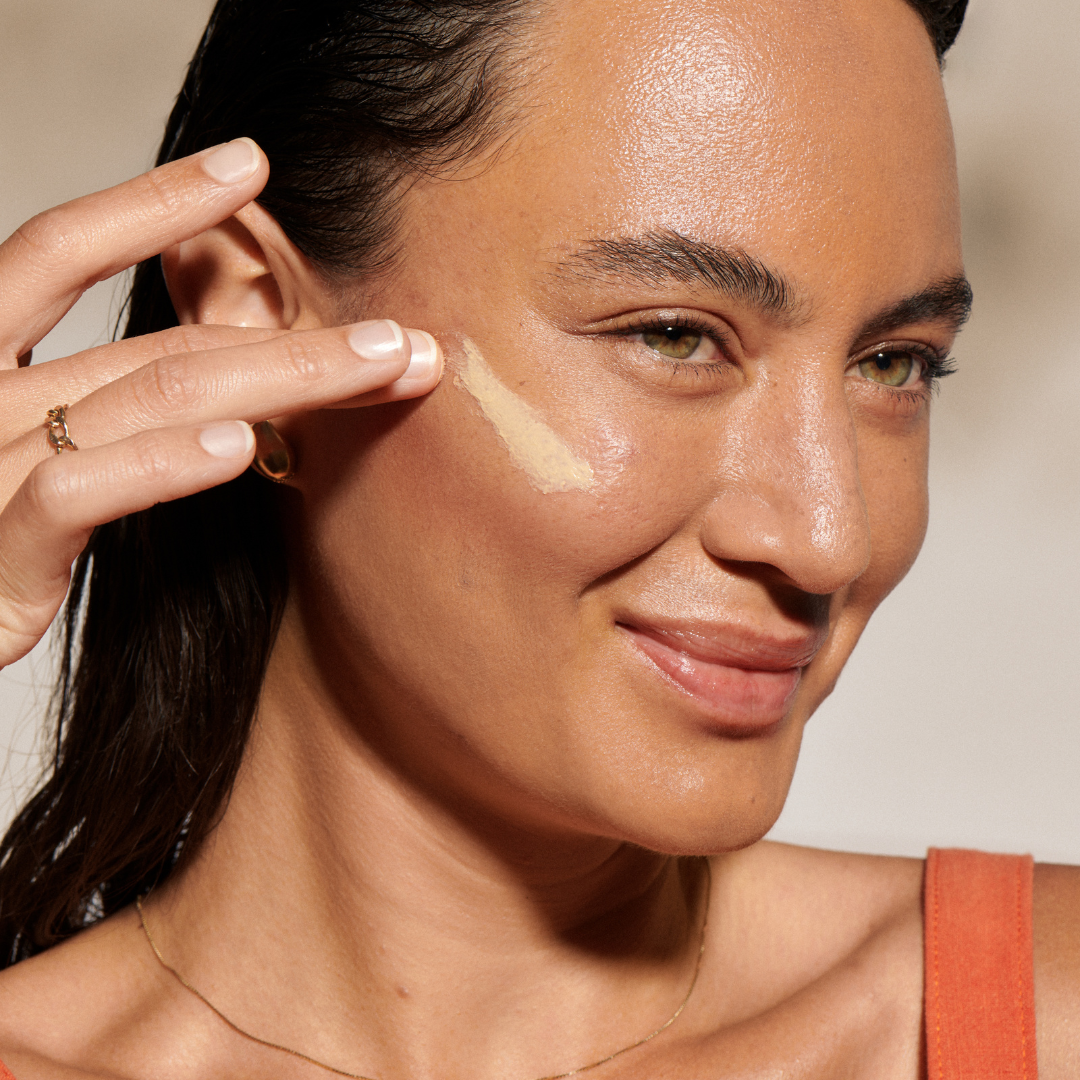 A woman with glowing skin smiles as she applies a cream product to her cheekbone with her fingers.