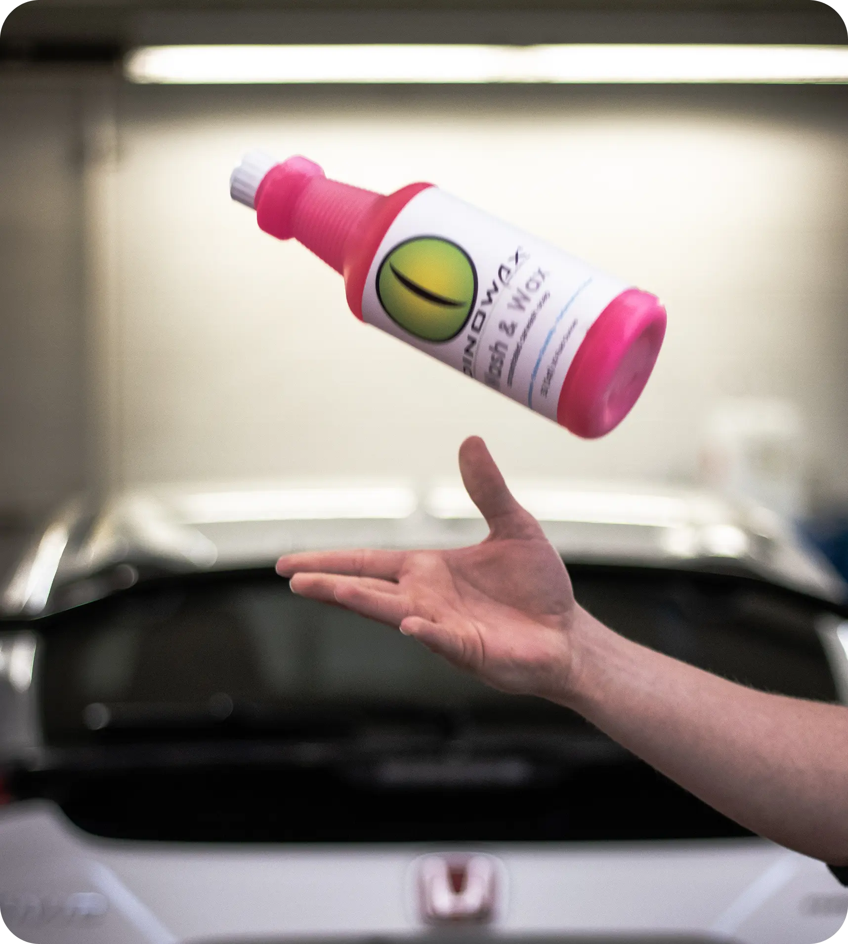 A hand tossing a pink bottle labeled 'Wash & Wax' in front of a car.