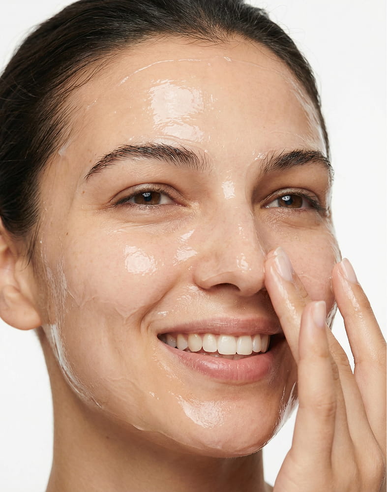 A close-up of a smiling woman applying a clear gel skincare product to her face.