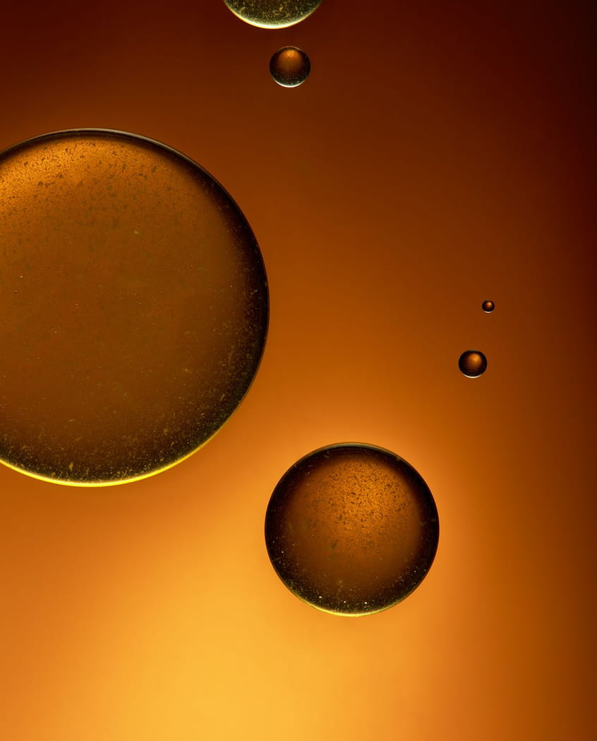 A macro photograph of golden oil droplets of various sizes against a warm, amber-colored gradient background.