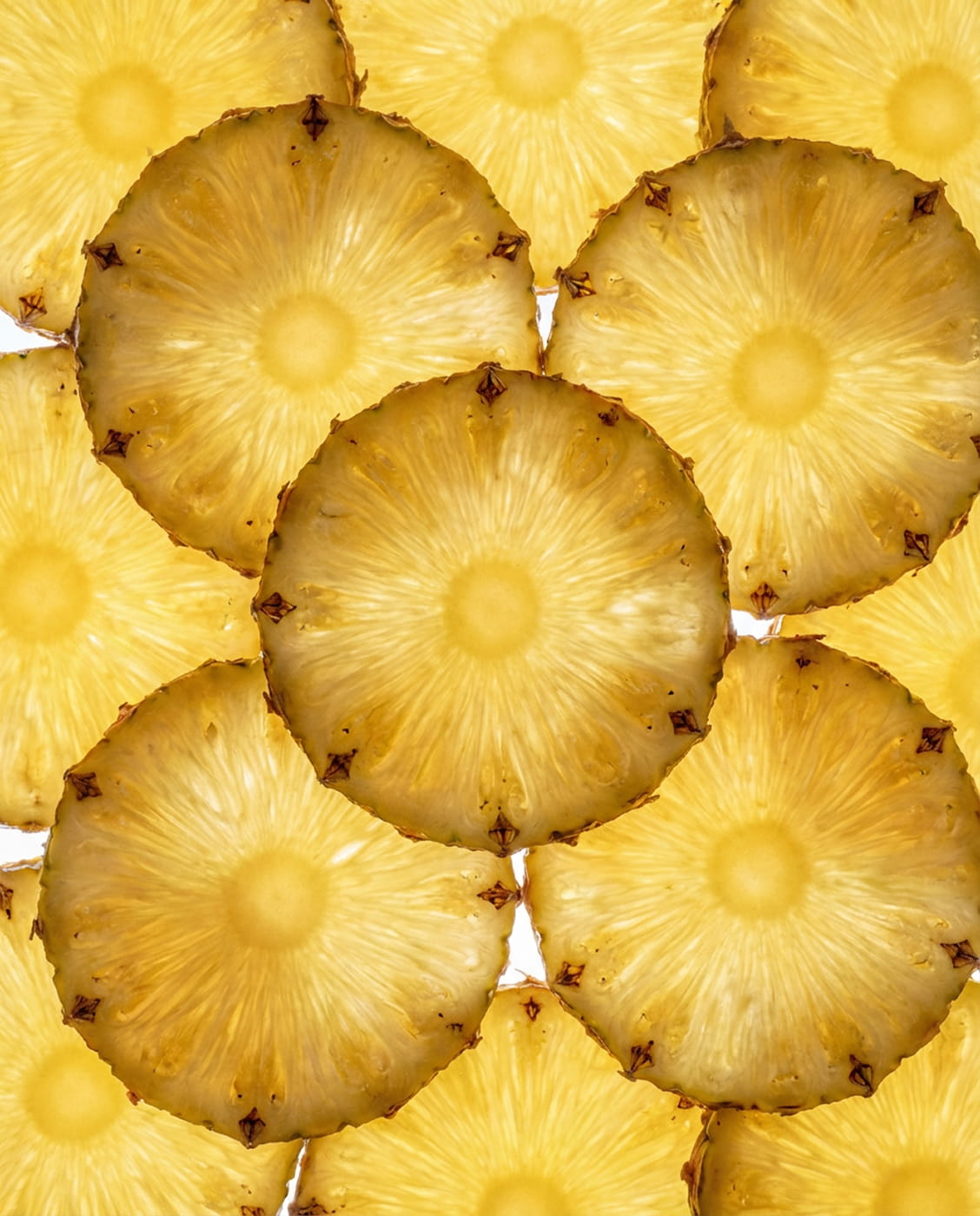 A close-up, backlit shot of several overlapping, round slices of pineapple against a white background.