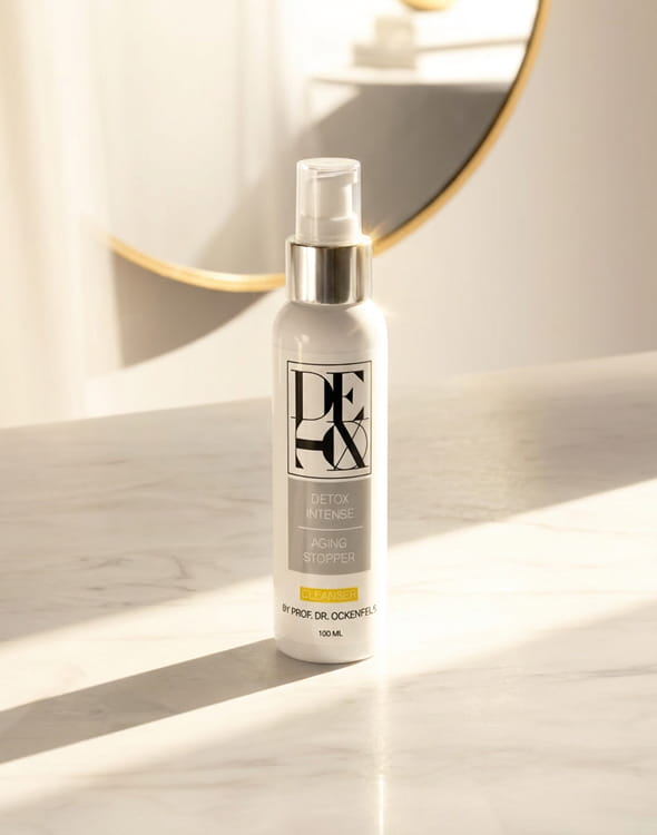 A white bottle of DE|OX Intense Cleanser standing on a marble surface in front of a round mirror.