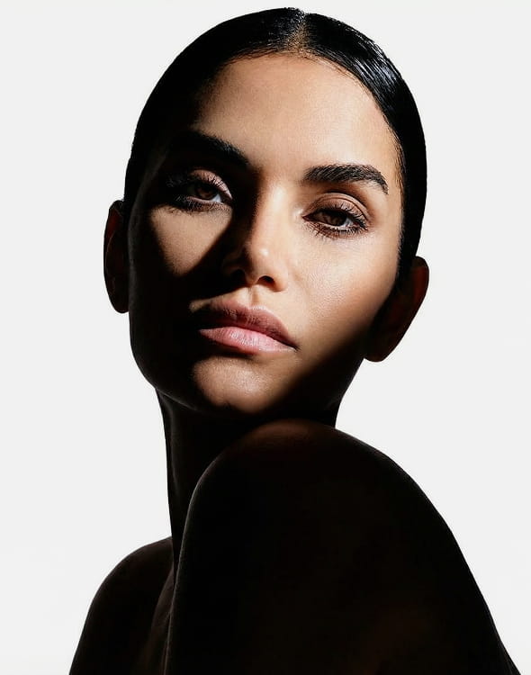 A close-up portrait of a woman with slicked-back dark hair, her face partially obscured by shadow.